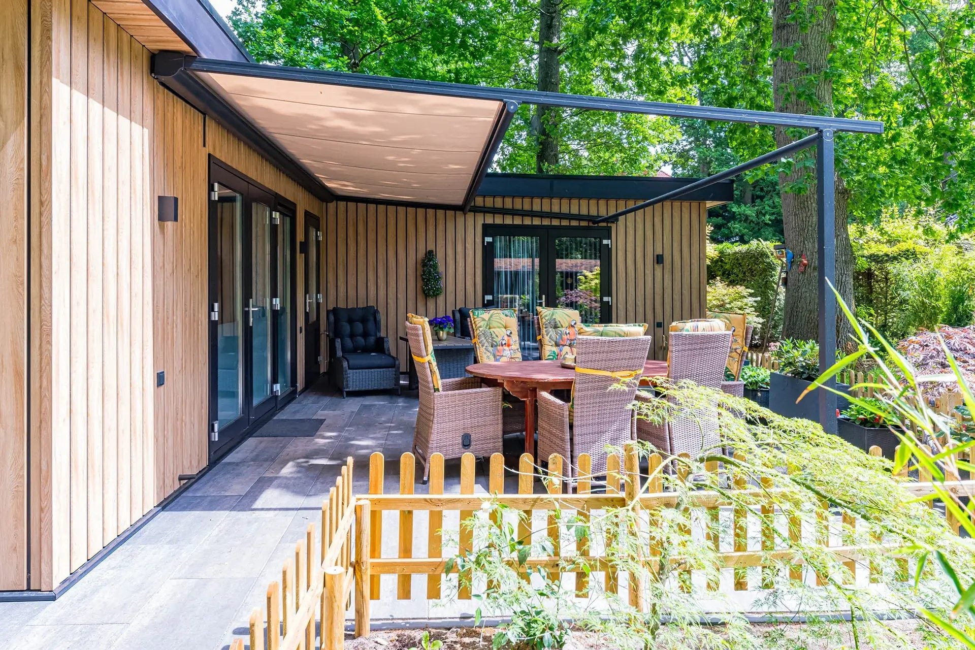 Antraciet pergola met uitgerold beige doek boven een terras met eettafel, tegen een woning met houten gevel