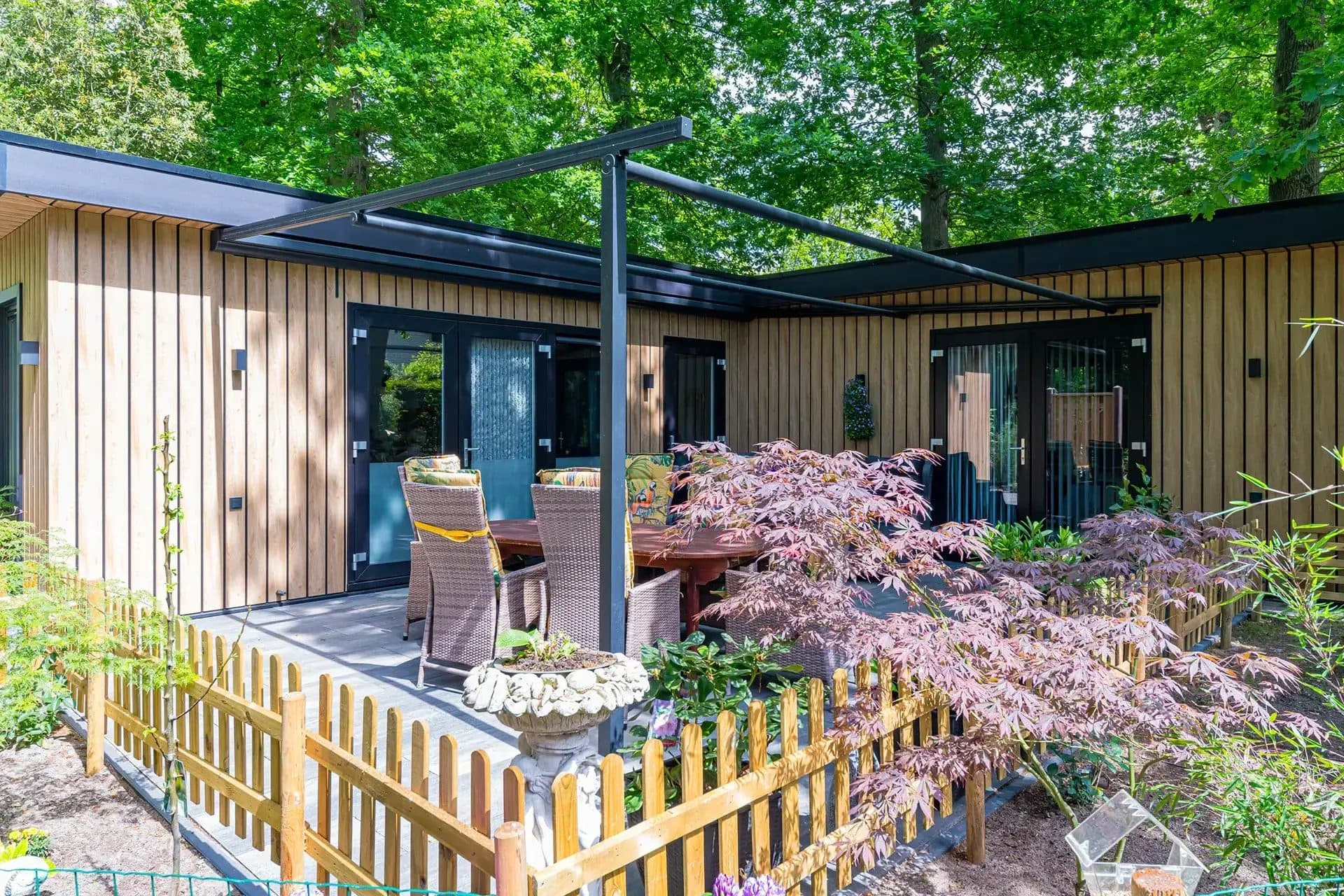 Antraciet pergola met opgerold doek naast een woning met houten gevel, met tuinset en een Japanse esdoorn