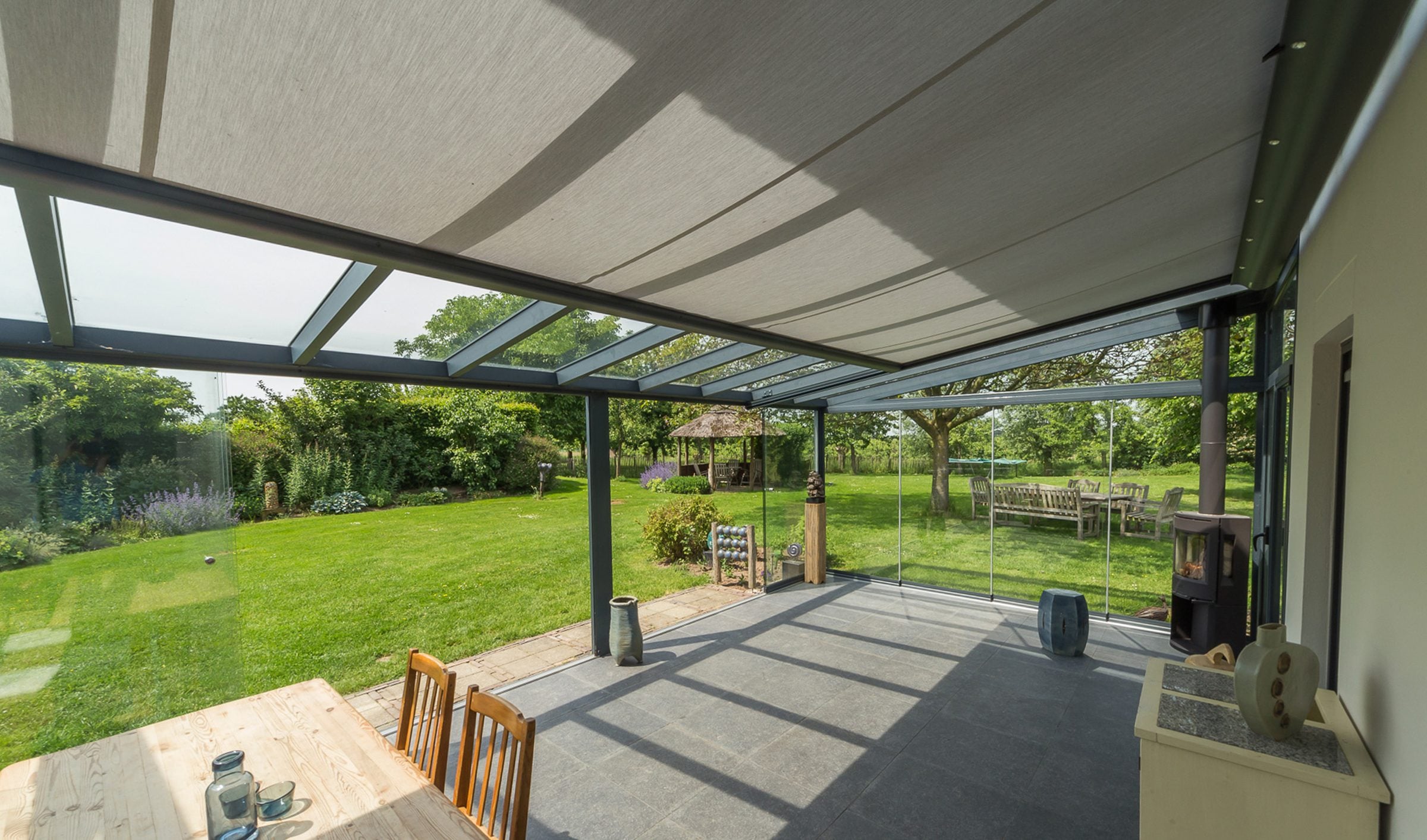 Zicht van onder een glasdak met lichtgrijze onderdakzonwering over een houten tafel naar een grote tuin met fruitbomen