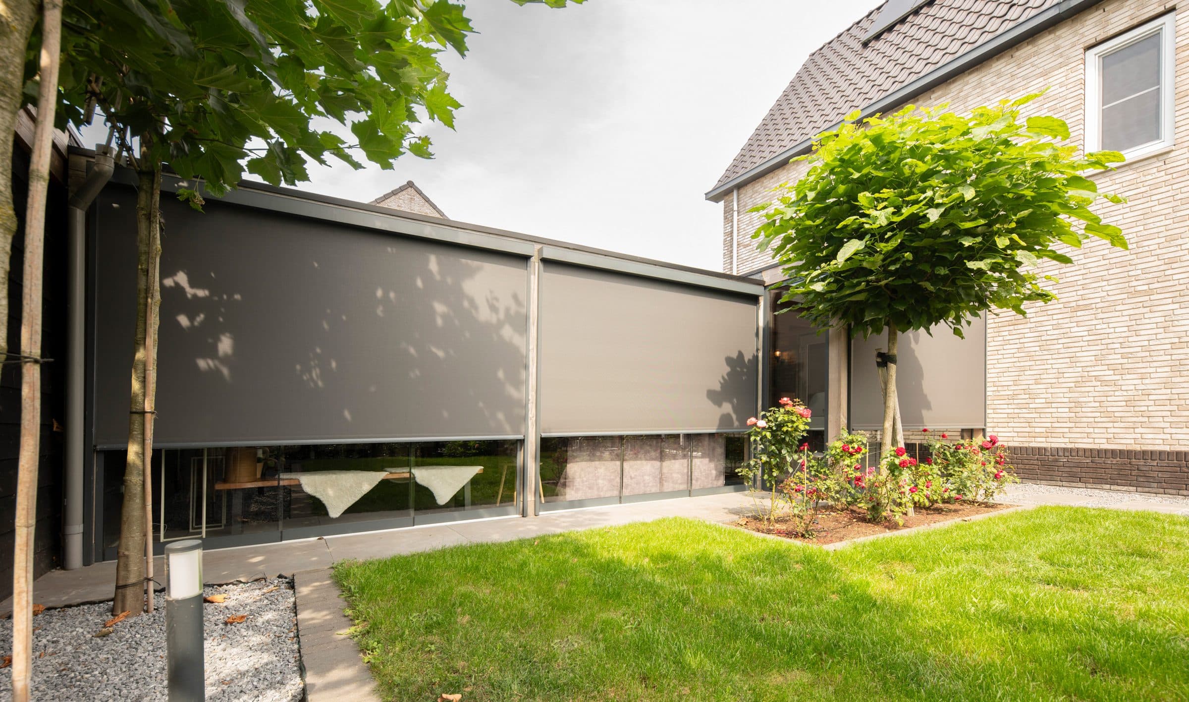 Neergelaten grijze screens over de hele achtergevel van een woning, met een gazon en bloeiende rozen