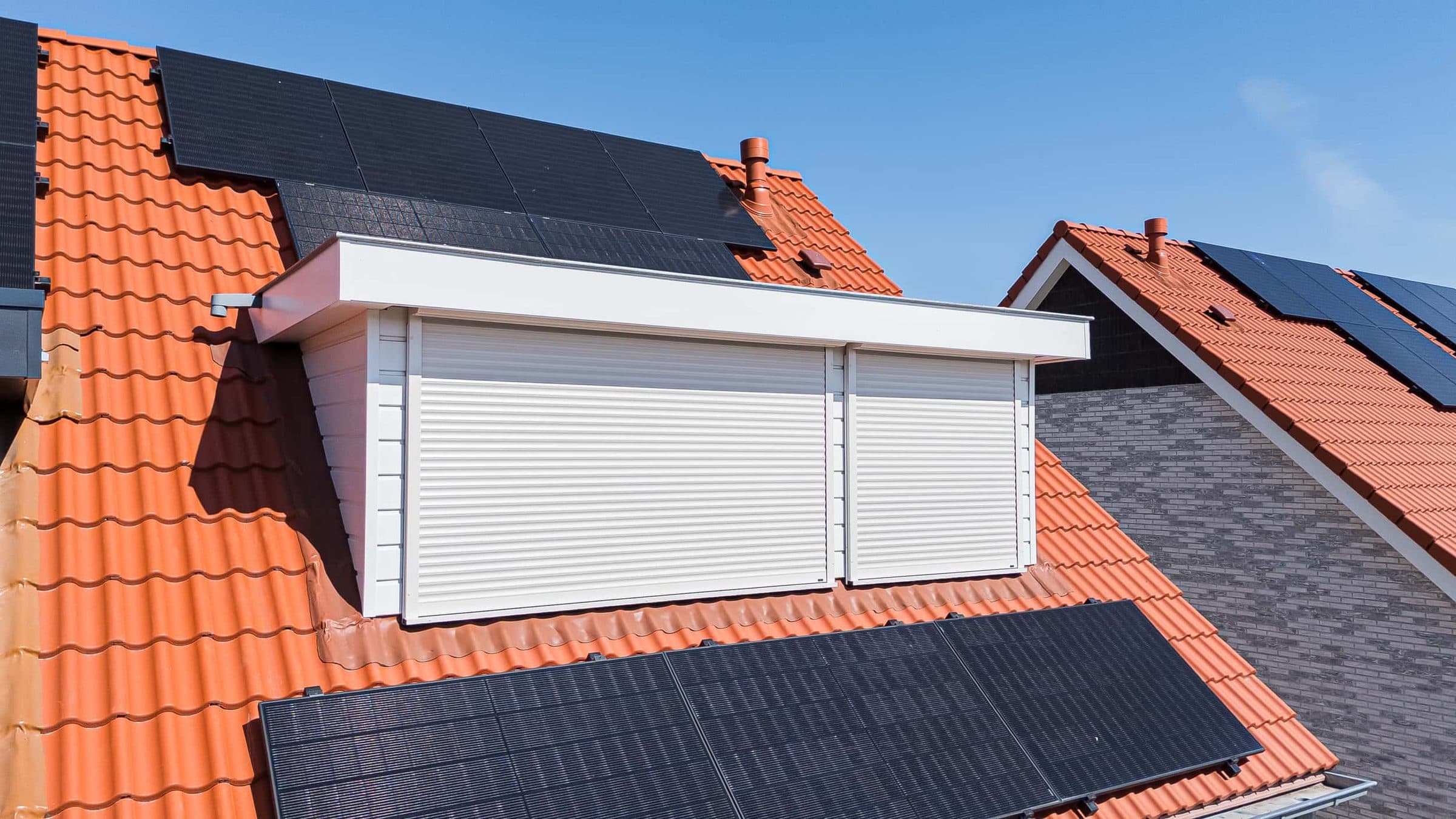 Witte dakkapel met twee gesloten witte rolluiken op een oranje pannendak met zonnepanelen