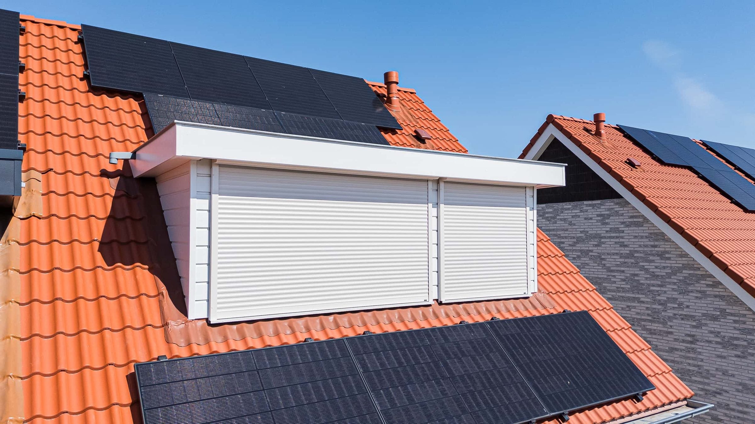 Witte dakkapel met twee gesloten witte rolluiken op een oranje pannendak met zonnepanelen