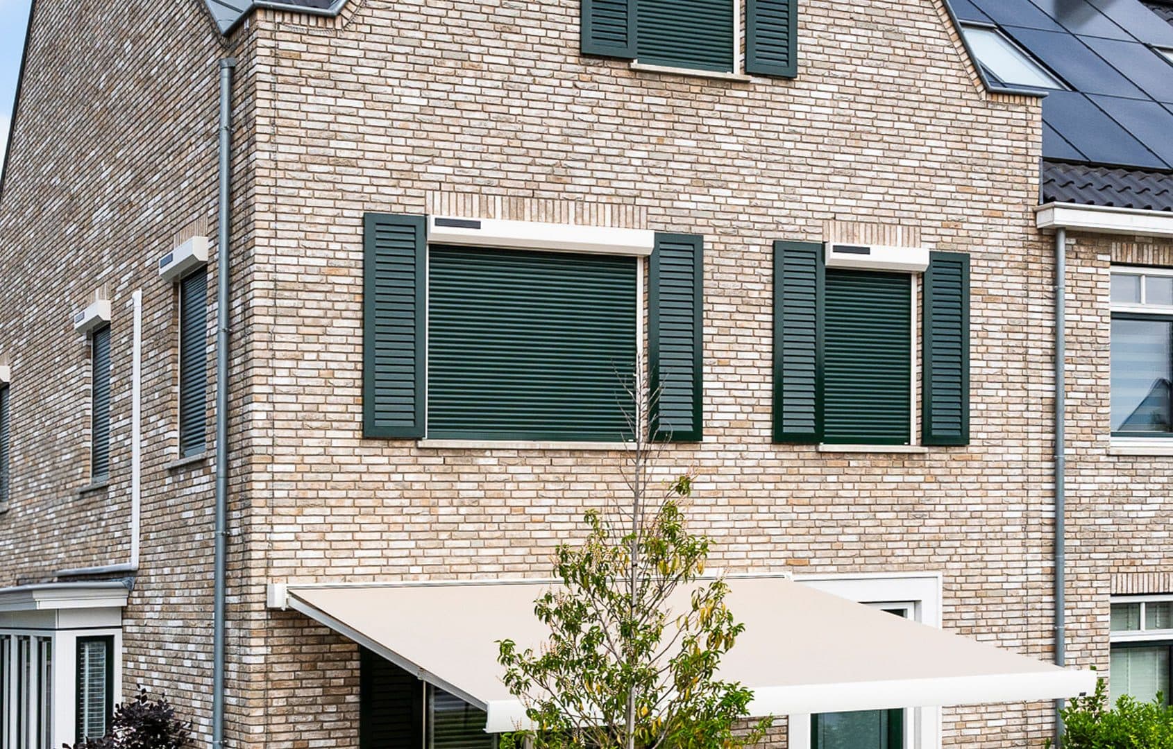 Groene rolluiken en luiken op een lichte bakstenen gevel, met een uitgerold knikarmscherm boven het terras